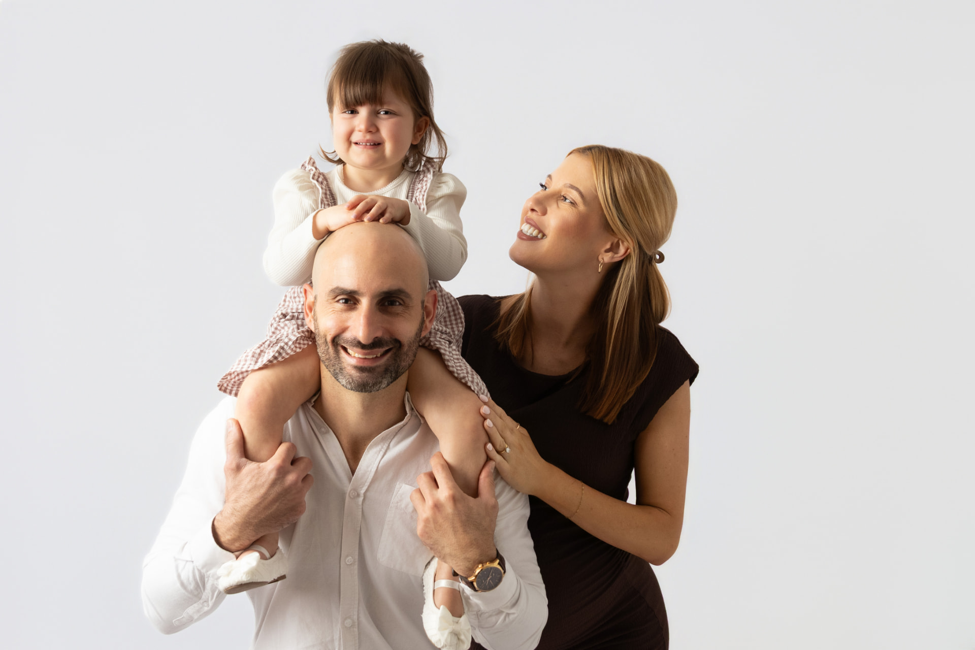 Toddler sitting on her dad's shoulders while her mum smiles beside them on a bright white backdrop