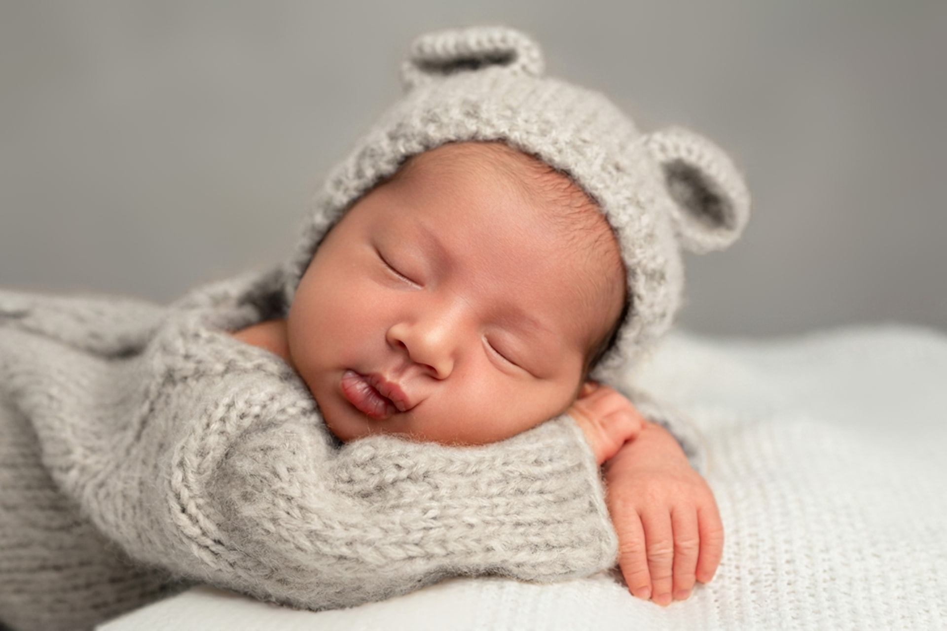 Sleeping newborn in a knitted grey bear bonnet resting on soft white blankets