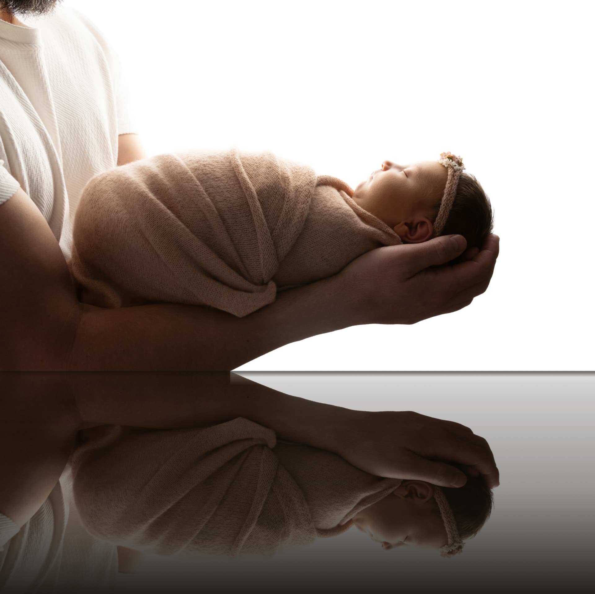 Newborn baby wrapped in soft beige fabric, held in cupped hands above a mirrored surface